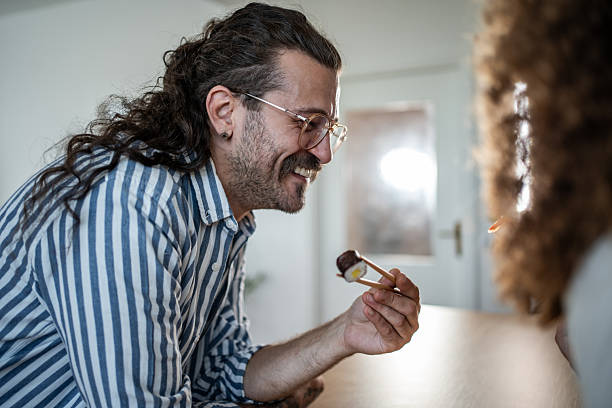 Smiling man with glasses and a striped shirt using chopsticks to eat sushi, sharing a happy moment with an unseen person, symbolizing enjoyment, dining, and casual dates