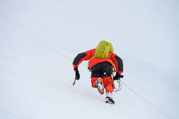Professional mountaineer dressed in high-altitude gear, including a bright jacket and heavy backpack, ascends a steep, snowy slope in the high mountains. The challenging conditions demand intense focus and determination as they navigate the harsh environment.