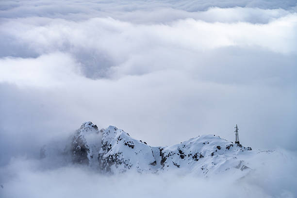 A communication tower stands prominently on a snowy mountain peak, enveloped in dense fog. The mist creates an ethereal atmosphere, obscuring the surrounding landscape and highlighting the tower's solitary presence at high altitude.