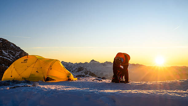 Experienced mountaineer organizing equipment beside a bright yellow tent as the sun rises over the snowy High Mountain base camp, setting a determined and focused atmosphere.