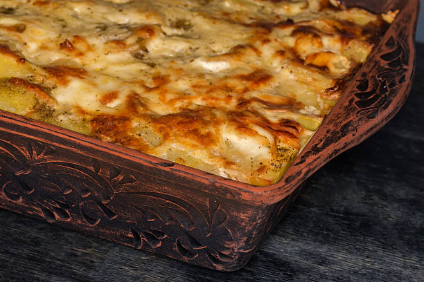 Baked potato casserole with browned crust of the melted cheese on top in the rectangular clay oven tray on a black wooden table, fragment close-up