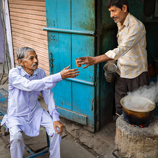A street-side tea vendor hands a small clay cup of steaming chai to an older seated customer outside a weathered turquoise shop door. A pot boils over an open stove as the vendor holds a metal kettle and strainer, while the seated man reaches out to accept the cup. The scene captures a quiet, everyday exchange at a local tea stall with warm, worn colors and visible steam.