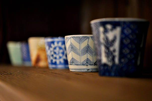Small ceramic cups with various intricate patterns are arranged in a row on a wooden surface. Each cup features unique designs, predominantly in blue and white tones. The focus highlights the craftsmanship of the glaze and patterns, commonly seen in traditional Japanese ceramics. The perspective of the image creates a depth effect among the cups.