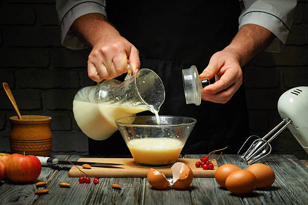 A chef pours milk from a glass jug into a bowl containing eggs and other ingredients. The kitchen is dimly lit, creating a warm ambiance as fresh apples and eggs are nearby