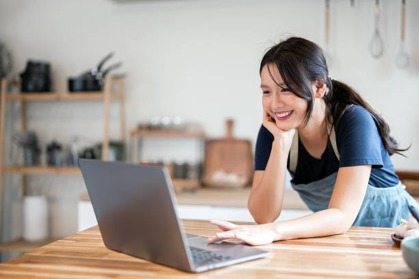 istock Cheerful Asian woman in an apron smiling and casually interacting with her laptop while resting her chin on her hand in the kitchen. 2247282286