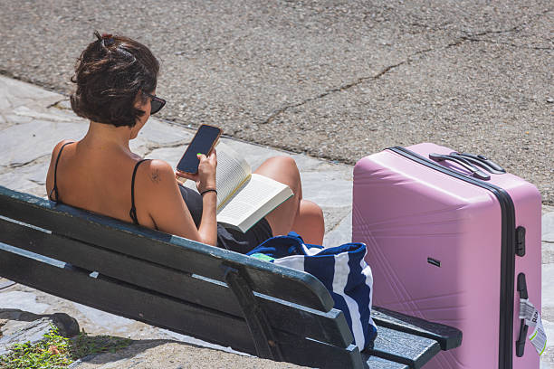 Cadaques, Spain - September 7, 2024: woman with a suitcase waits on a bench while reading a book