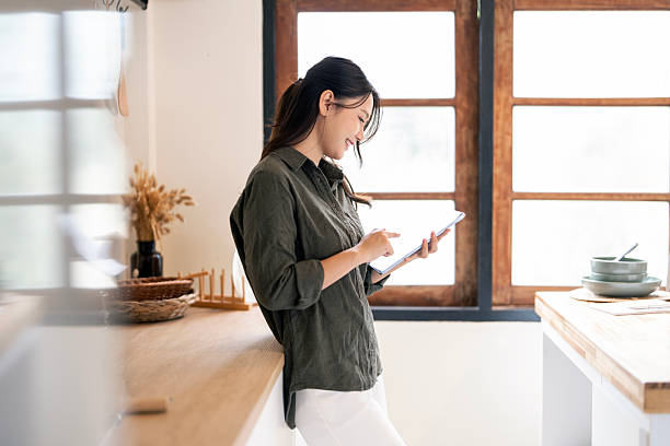 istock Young Asian woman leaning on a kitchen counter and happily using a digital tablet for work or communication. 2247282925