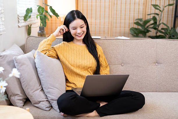 istock Smiling young Asian woman sitting cross-legged on a sofa, happily working or browsing on her laptop in a bright, plant-filled living room. 2247284282