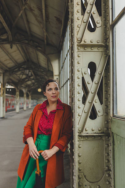 Adult woman in rust-orange coat and green trousers stands leaning against riveted metal support column on old, covered train station platform, holding umbrella