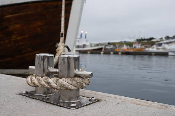 Close-up of a metallic cleat with rope on a concrete dock, featuring a blurred wooden boat and marina in the background, evoking a peaceful maritime scene