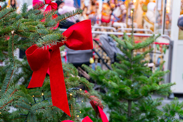 istock Christmas tree with a large red bow at a Christmas market, featuring a blurred carousel and festive decorations in the colorful background. 2247296402