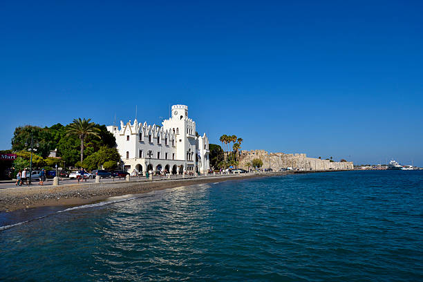 Kos, Greece - September 16, 2025:  The imposing police headquarters building on Akti Miaouli Street with a separated bike lane and pedestrian walkway along the riverbank. In the background, part of Neratzia Castle.