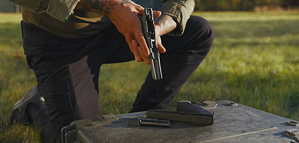 Trained soldier with hand tattoos performing the disassembly and the cleaning process of a pistol gun firearm in field conditions. Professional technician removing parts to clean and perform maintenance