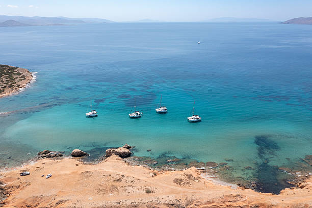 Four catamarans float peacefully in shallow blue waters near the rocky coast of Paros, Greece. The vivid sea contrasts with the dry, rugged land along the shoreline.