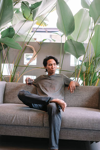 istock Thoughtful Young Asian Man Relaxing on Sofa with Drink 2247310991