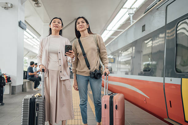 Two Asian female travelers with luggage walk along the platform at a train station, searching for their train.