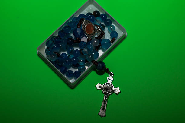 A Catholic rosary with blue agate beads and a metal crucifix is neatly folded inside a clear plastic box, alongside a silver ring with a stone. This religious set is placed on a contrasting, bright green background, symbolizing faith, prayer, and spiritual serenity.