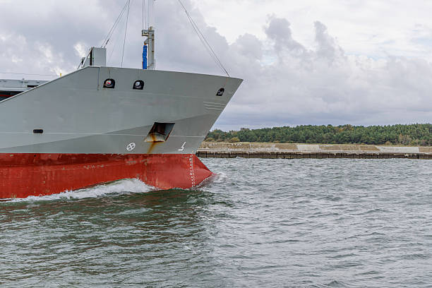 The grey bow and red hull of a large vessel slice through the choppy water, creating a wake near a concrete and forested shoreline under a cloudy sky.