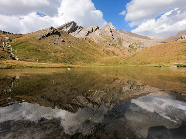 Peaceful alpine landscape in the French Alps featuring a clear mountain lake with mirror-like reflections of rugged peaks and a partly cloudy sky. The scene captures the serene beauty of nature, with earthy tones, grassy slopes, and tranquil water, ideal for travel, nature, and outdoor themes.