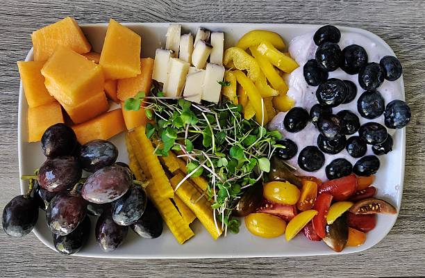 Delicious fresh food, including cantaloupe melon, BellaVitano Merlot cheese, yellow bell pepper, blueberry yogurt with blueberries, cherry tomatoes, microgreens, yellow carrot, purple grapes