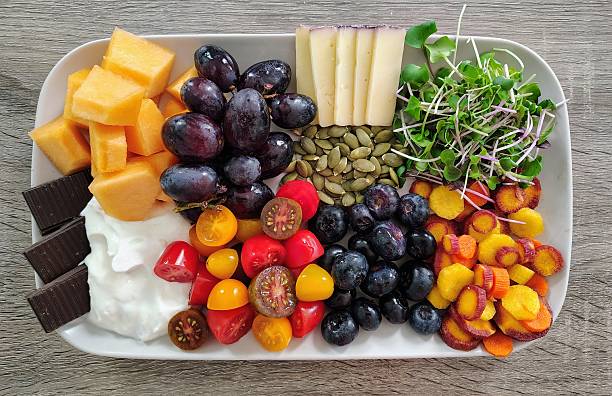 Delicious fresh food, including yellow, orange, and purple carrots, blueberries, cherry tomatoes, yogurt, dark chocolate, cantaloupe, purple grapes, pumpkin seeds, BellaVitano Merlot cheese, microgreens