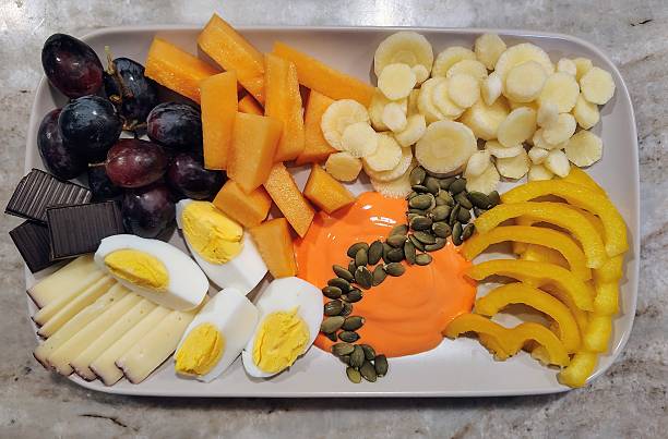 Delicious fresh food, including white carrots, cantaloupe, purple grapes, dark chocolate, egg, cheese, mango yogurt, pumpkin seeds, yellow bell pepper