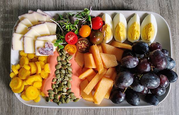 Delicious fresh food, including yellow carrots, mango yogurt, pumpkin seeds, cantaloupe, purple grapes, hard boiled eggs, microgreens over mixed baby greens, cherry tomatoes, BellaVitano Merlot cheese, raspberries
