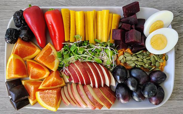 Delicious fresh food, including oranges, apple, Ajwa dates, dark chocolate, pumpkin seeds, yam, microgreens over hummus, purple grapes, egg, beets, yellow carrot, red bell peppers