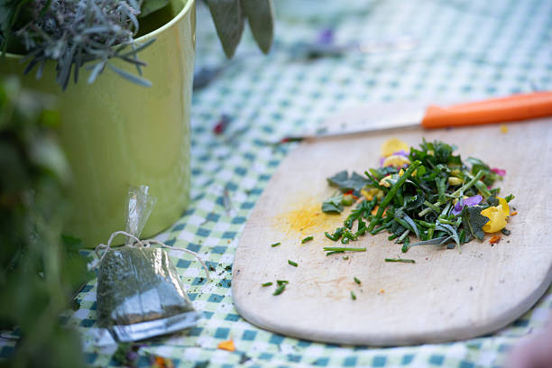 A close-up of a wooden cutting board with freshly chopped herbs and edible flowers, next to a knife with an orange handle. A green plant pot and a small bag of herbs are visible on a checkered tablecloth, highlighting the preparation of natural ingredients.