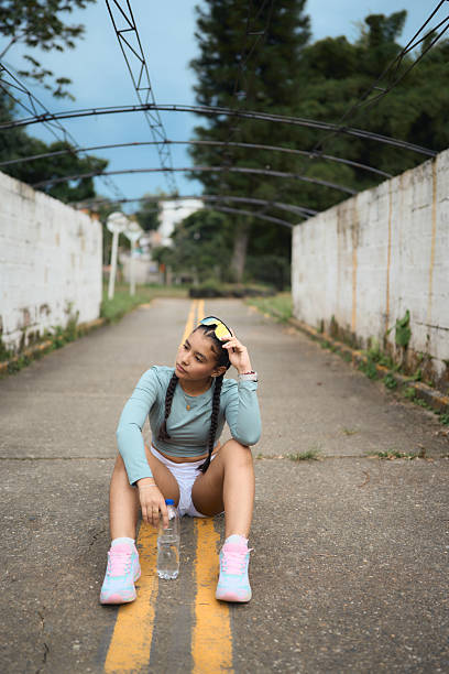 Young athletic woman with braids pausing for a break after exercising outdoors, hydrating with a water bottle on a road