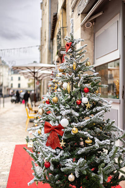 istock Festive Christmas tree with white artificial snow and large red bows stands on a red carpet near a cafe entrance on a European city street in winter 2247336245