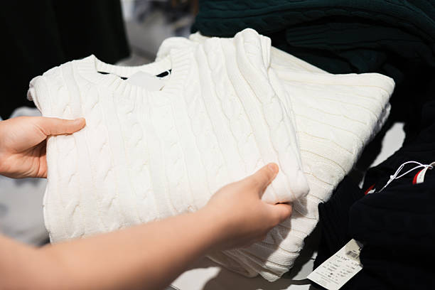 istock A person's hands hold a folded white cable knit sweater in a clothing store, with other garments visible nearby. 2247336793