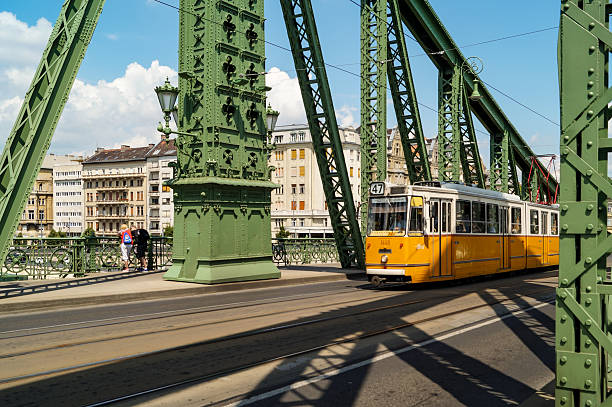 The classic yellow tram (line 47) is seen crossing the green, monumental truss structure of Budapest's Liberty Bridge. The cityscape of buildings is visible on the Pest side, providing an urban background to the scene.