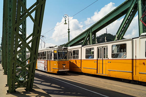 A classic yellow tram with line number 49 is passing over the green steel structure of the Liberty Bridge in Budapest, Hungary. The iconic Ganz UV tram operates on a sunny summer day, showcasing European public transportation.