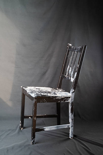 Paint spattered wooden chair under spotlight creating nostalgic studio mood worn seat and peeling finish show layers of white paint and past projects solitary