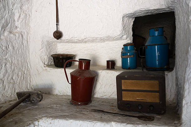 A rustic kitchen shelf built into textured white walls, displaying colorful vintage metal containers, a red enamel pitcher, and an old radio. The worn surfaces and traditional utensils evoke a nostalgic rural atmosphere.
