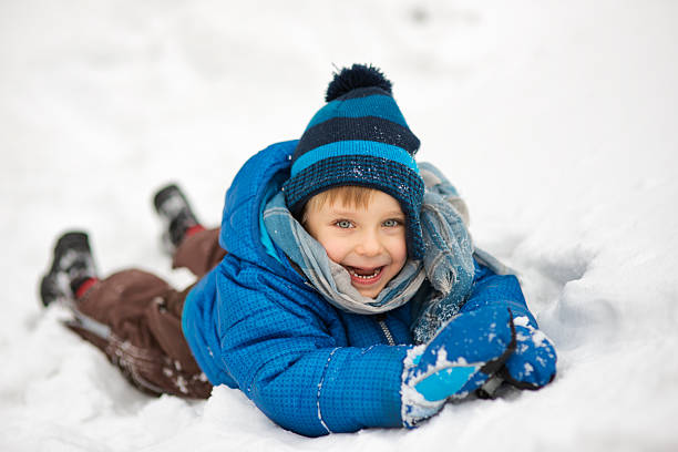 A cute laughing child dressed in a blue winter jacket, brown snow pants, and a striped knit hat with a pom-pom lies joyfully in the snow. Wearing a matching scarf and gloves, the boy smiles brightly, surrounded by a blanket of white snow. Shot with Nikon D800.
