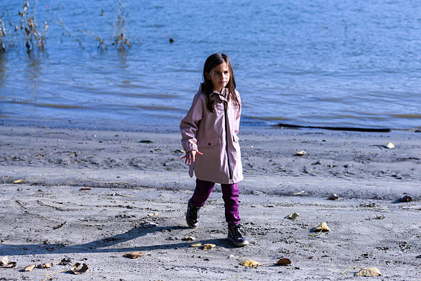 Young girl walking alone along a sandy river bank in autumn, wearing a pink coat, looking thoughtful and reflective