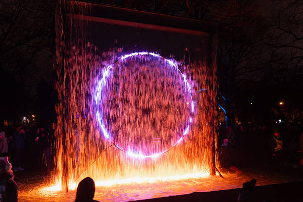 Waterfall curtain lit by vivid circular light ring producing dynamic mix of colors and reflections during outdoor evening event with people watching illuminated artistic installation