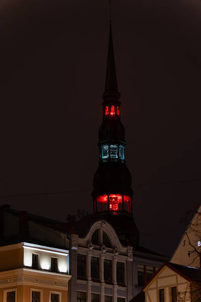 Tall historic tower in Riga illuminated with red and blue lights creating dramatic nighttime architectural scene with surrounding rooftops and ornate facade details