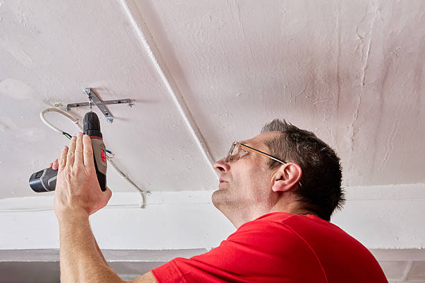 istock Fixing electrician uses cordless screwdriver to secure ceiling bracket for LED chandelier in rural house, wires held close to mounting area. 2247358002