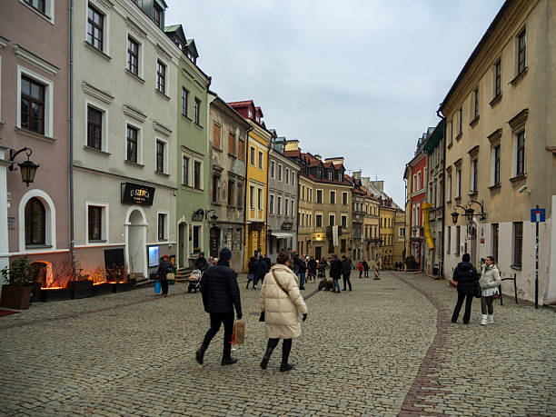 A wide cobblestone street in Lublin's Old Town is lined with colorful, historic buildings sloping towards the center. People are seen walking in the foreground and down the street on a cool, overcast day.