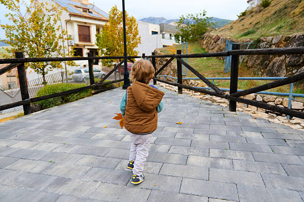 A toddler in a brown puffer vest walks along a gray paved walkway in a park during fall. Leaves on the ground, wooden fence, hillside and trees in autumn colors.