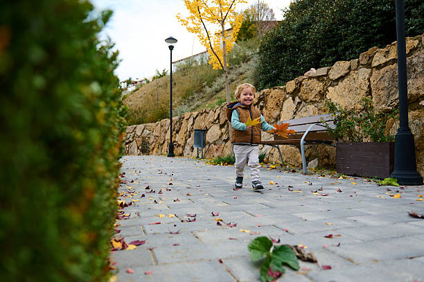 A happy toddler in a brown vest and light jacket plays with autumn leaves along a park path, showing warmth, care, and togetherness with mom on a sunny fall day.