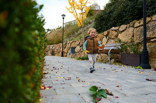 A cheerful toddler in a brown vest explores an autumn park, scattering leaves along a stone walkway beside a bench and lampposts.