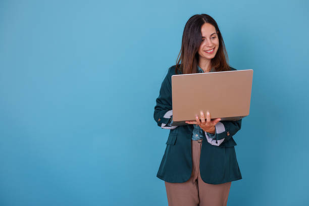 istock Smiling young woman is standing and happily engaging with a laptop against a blue backdrop 2247366162