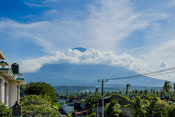 A scenic photograph of the volcano near Amed, Bali, captured from afar with lush tropical forest vegetation in the foreground. The volcano’s peak rises into the clouds under a bright blue sky, highlighting the dramatic landscape and natural beauty of the region.