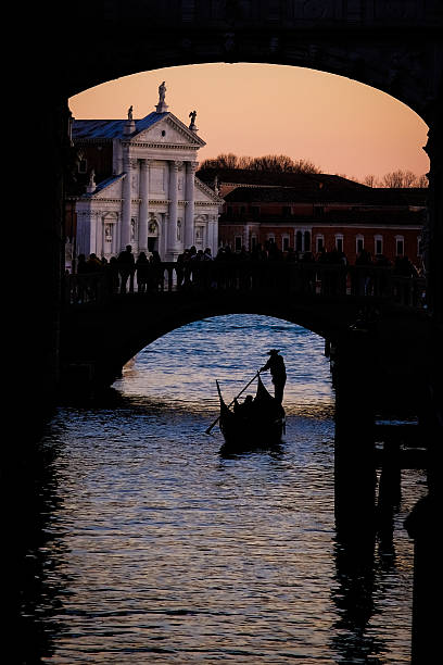 A Venetian gondolier guides his gondola through a busy canal at sunset, framed by arching bridges and historic architecture. Warm evening light reflects off the water, capturing the timeless charm and atmosphere of Venice.
