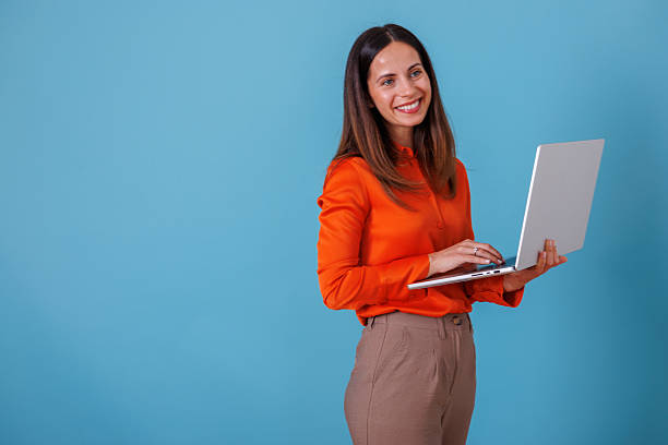 istock Professional woman smiling, working on laptop, standing against a blue background, representing remote work and business technology 2247371127