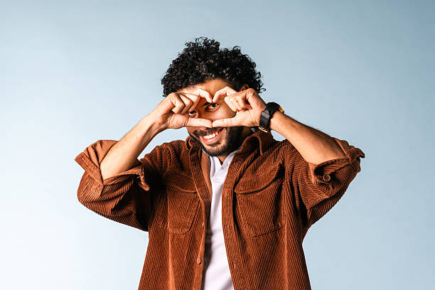 A cheerful middle-aged Middle Eastern man in a casual brown shirt makes a heart gesture with his hands on a soft blue background.
